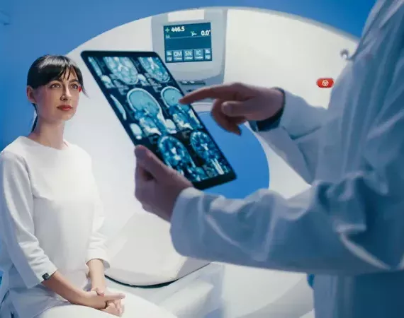 Frau sitzt neben MRT-Gerät und unkenntlicher Arzt im Vordergrund begutachtet einen Gehirn-Scan.