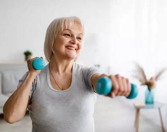 Ältere Frau trainiert zuhause lachend mit Hanteln.
