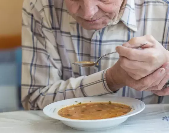 Parkinsonkranker Senior führt beim Suppe-Essen seine Hand, die den Löffel hält, mit der anderen.