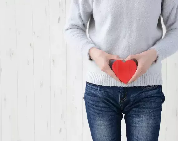 Unkenntliche Person in Pullover und Jeans hält ein rotes Herz in Höhe des Darms vor den Körper.