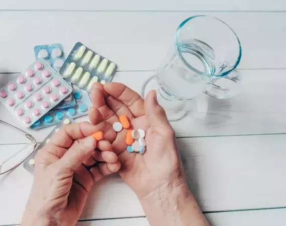 Alte Hände mit vielen Tabletten und Kapseln, Medikamenten-Blister und Wasserglas auf dem Tisch.